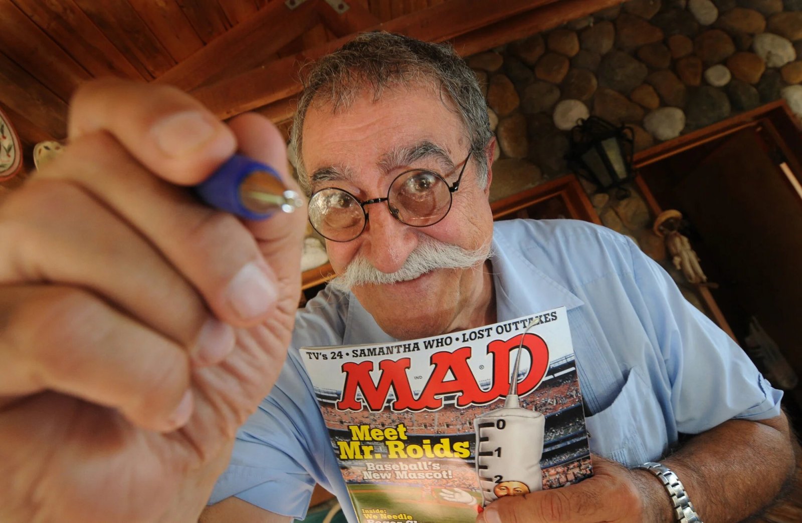 A man with a white mustache and round glasses holds up a copy of MAD magazine and points a pen toward the camera