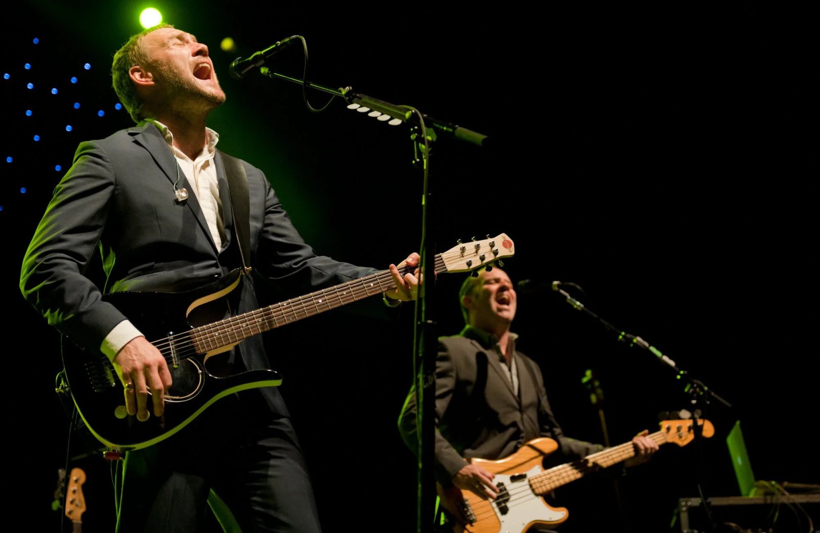 Singer David Gray sings into a microphone with his guitar as his bassist plays behind him