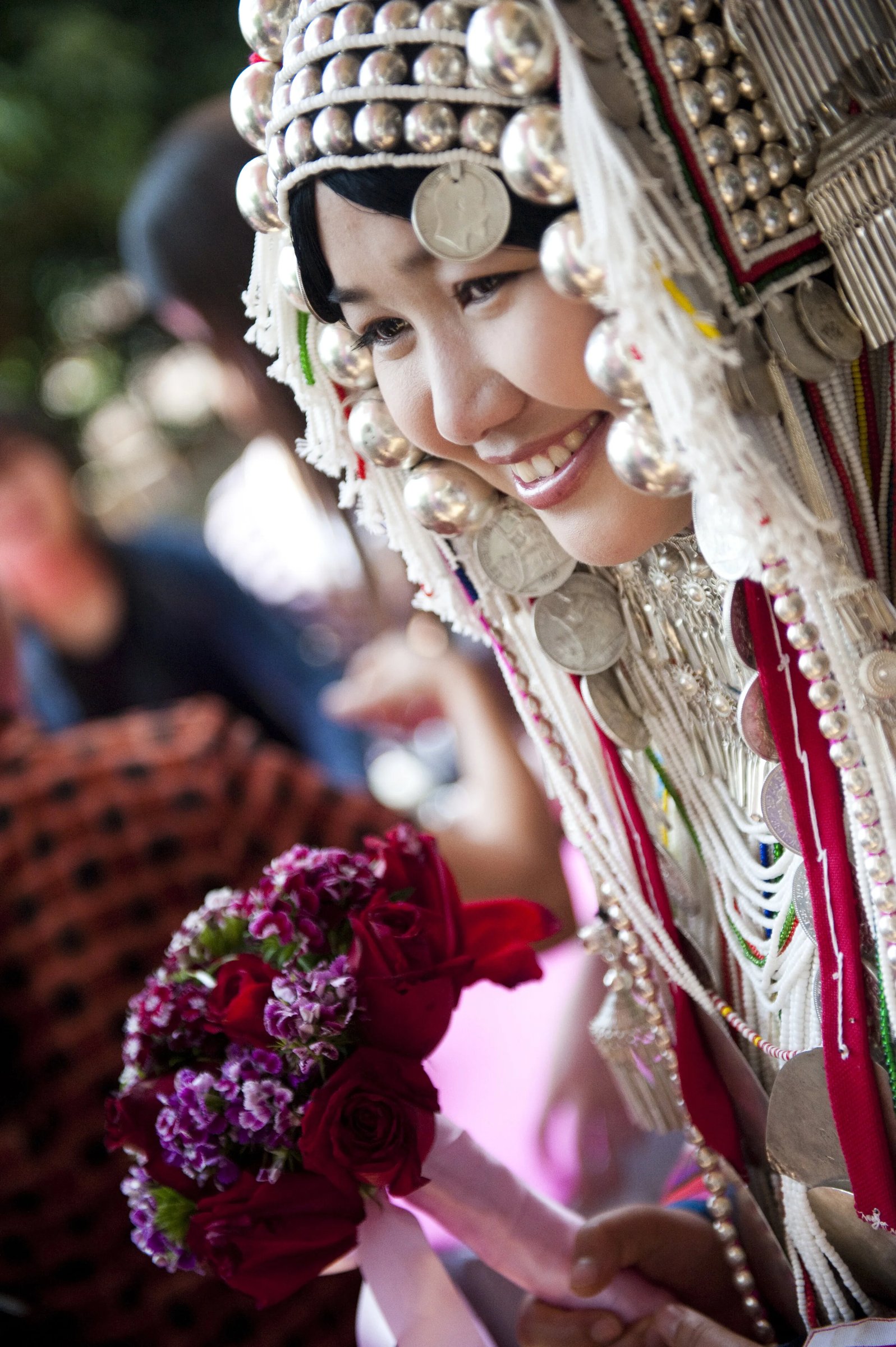 A smiling woman in a traditional silver headdress holds a bouquet of flowers in Chiang Rai, Thailand