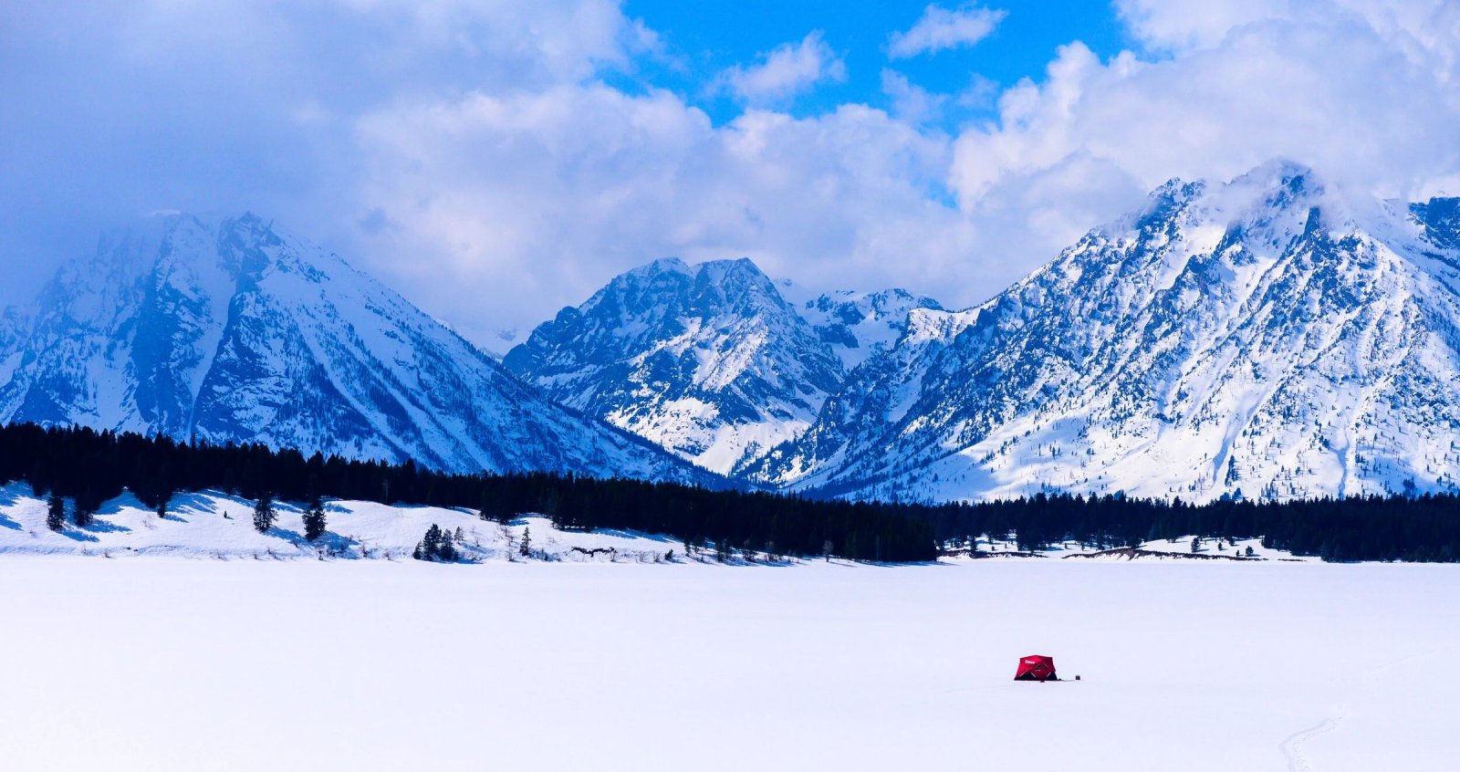 Snow-covered mountain peaks rise above a wide, snowy plain under a blue sky